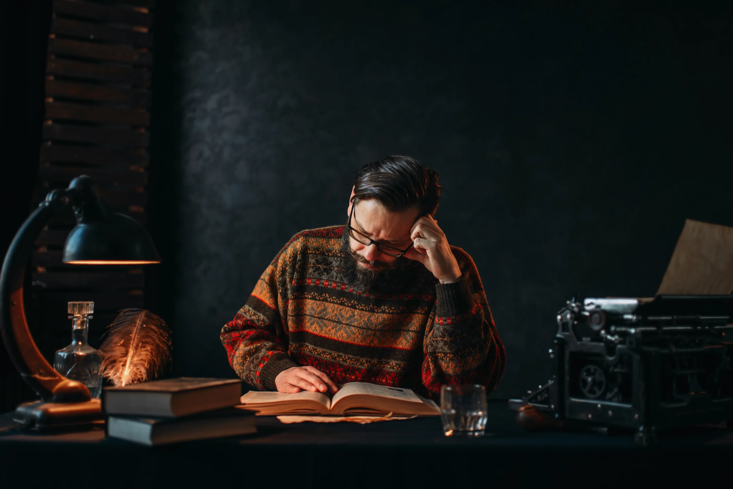 Man in a cozy sweater reads intently at a dimly lit wooden desk, surrounded by books, a lamp, a feather quill, and an old typewriter; thoughtful mood spineauthors