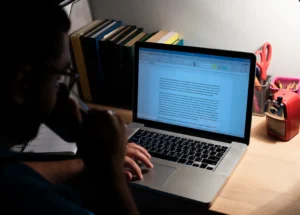 A person working at a desk lit by a lamp, typing on a laptop. Books and stationery are nearby, creating a focused and studious atmosphere spineauthors
