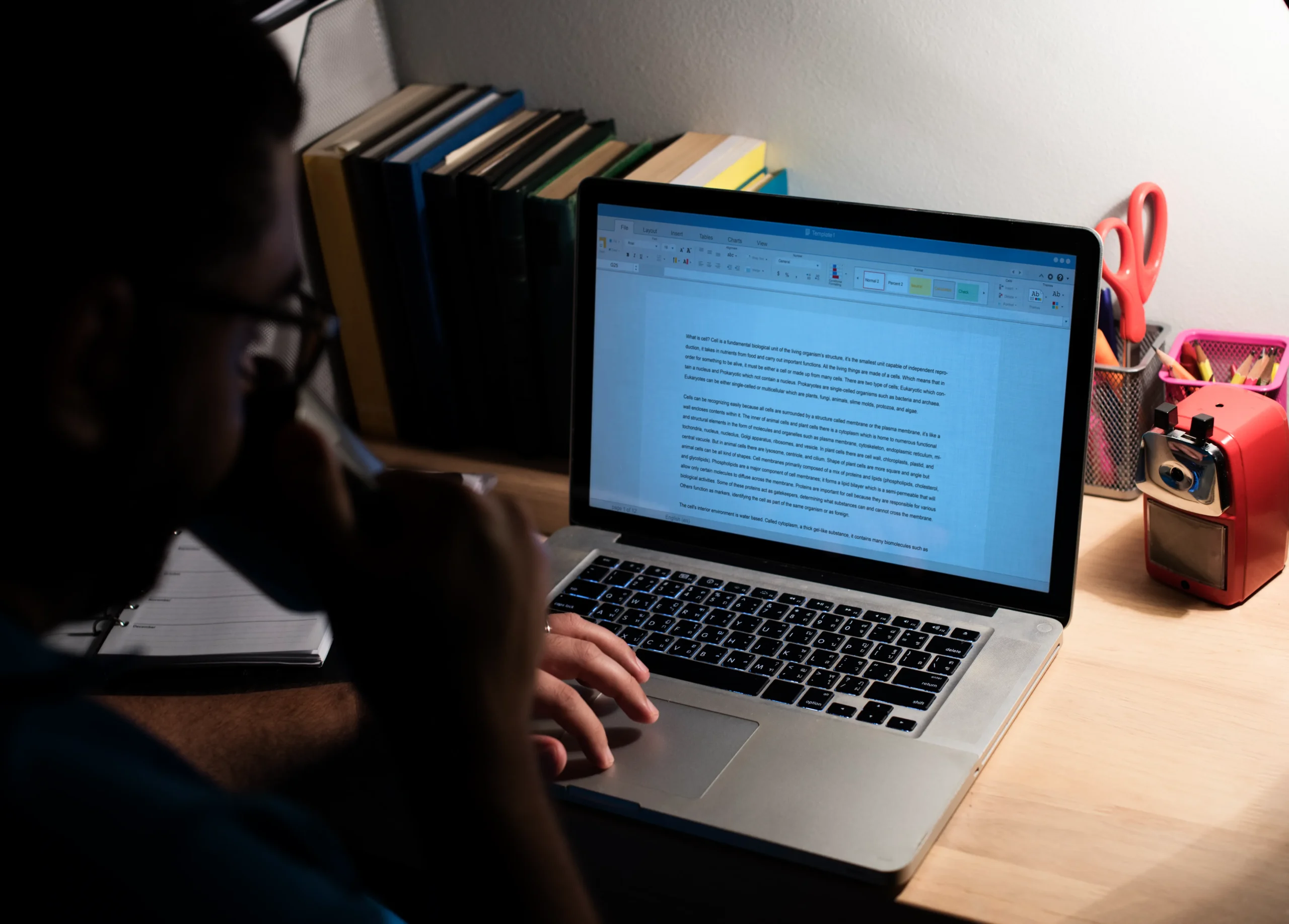 A person working at a desk lit by a lamp, typing on a laptop. Books and stationery are nearby, creating a focused and studious atmosphere spineauthors